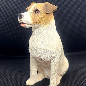 Puede incluir: Figura detallada de un perro Jack Russell Terrier sentado. El perro tiene un pelaje blanco con marcas marrones en la cabeza y las orejas. La figura está sobre un fondo oscuro.