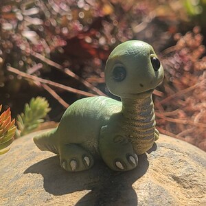 Può includere: Una piccola statuina di dinosauro verde con occhi grandi e un collo testurizzato. Il dinosauro è seduto su una roccia.