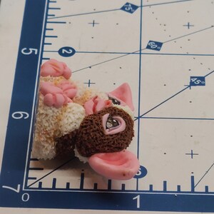 May include: A small, brown and white, furry animal figurine with pink ears and a pink nose. The figurine has a brown, textured body and a white, fluffy tail.