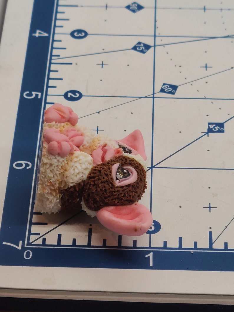 May include: A small, brown and white, furry animal figurine with pink ears and a pink nose. The figurine has a brown, textured body and a white, fluffy tail.