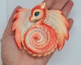 May include: A small, hand-crafted, red and white dragon figurine with a curled tail and wings. The dragon has a blue eye and is made of polymer clay.