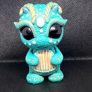 May include: A teal blue dragon figurine with gold accents and white spots. The dragon has large black eyes and is holding a brown and white striped object in its front paws.