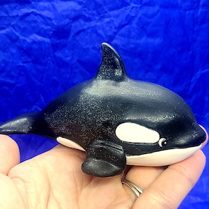 Op de afbeelding: Zwarte en witte orka-figuur met een glinsterende afwerking. De orka is in zwempositie met zijn rugvin omhoog en zijn staartvin omlaag.