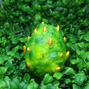 Pode incluir: Um objeto decorativo verde em forma de ovo com detalhes dourados em forma de cone. O objeto tem uma superfície texturizada e é cercado por folhagem verde. O tema geral sugere um item de fantasia ou inspirado na natureza.