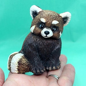 May include: A small, hand-painted ceramic figurine of a red panda sitting with its paws tucked in. The figurine is brown with white markings on its face and tail. The figurine is sitting on a person's hand.
