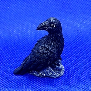 May include: A small black raven figurine perched on a grey rock base. The raven has detailed feathers and a realistic appearance.