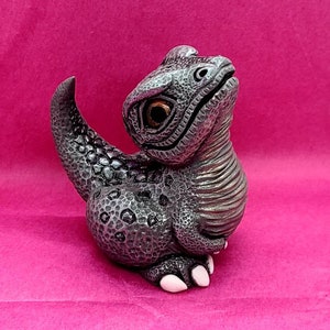 Pode incluir: Uma pequena figura de dinossauro prateado com uma superfície texturizada detalhada. A figura tem olhos grandes e expressivos com íris castanhos e unhas brancas. O dinossauro está em posição sentada, com a cauda enrolada.