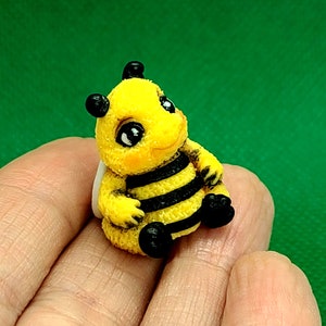 May include: A small, yellow and black bee figurine with large, black eyes and a friendly expression. The bee is sitting on a white surface.