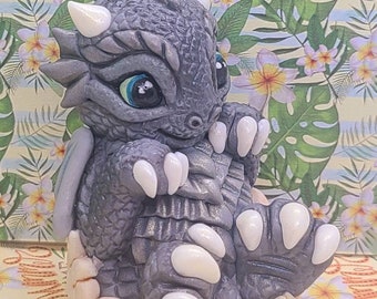 May include: A small, gray dragon figurine with white claws and blue eyes, sitting inside a light brown egg-shaped nest. The dragon has a textured, scaly body and is posed with its wings tucked in.