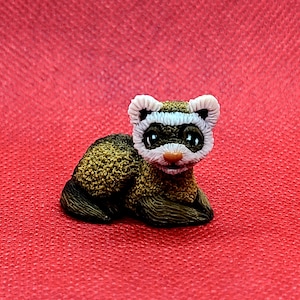 Könnte beinhalten: Kleine, detaillierte Figur eines Frettchens, liegend auf rotem Grund. Das Frettchen hat ein weißes Gesicht mit schwarzen Augen und einer braunen Nase. Sein Körper ist braun und beige, mit strukturierter Oberfläche. Eine Miniatur-Tierskulptur.