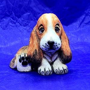 May include: A ceramic figurine of a brown and white basset hound puppy sitting with its paw raised. The puppy has a sweet expression and is painted realistically.