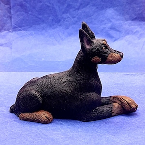 May include: A detailed figurine of a Doberman Pinscher dog, depicted in a relaxed pose. The dog's coat is black, with tan markings on its snout, chest, and paws. The figurine is set against a blue backdrop, showcasing the dog's realistic features and textured fur.