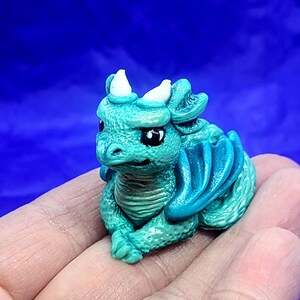 May include: A small, teal-colored dragon figurine with white horns and a blue wing. The dragon is sitting on its hind legs with its front paws tucked in.
