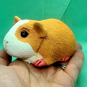 May include: A small, orange and white guinea pig figurine with a textured fur design. The figurine is made of a soft, pliable material and has pink feet.