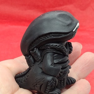 May include: A black and white figurine of an alien creature with a detailed, textured surface. The alien is in a crouching position with its arms crossed in front of it.