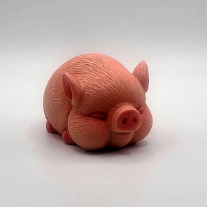 Op de afbeelding: Perzikroze keramische varkensbeeldje in slaappositie. Het varken heeft gesloten ogen, een rond lichaam en gedetailleerde gelaatstrekken. Het beeldje rust op een witte ondergrond.