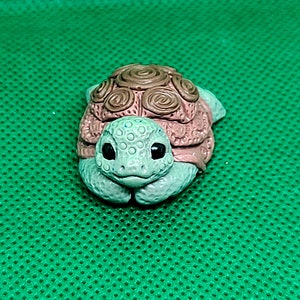 May include: A small, ceramic turtle figurine with a brown and pink shell and a green head. The turtle has a sweet expression and is sitting on a green surface.