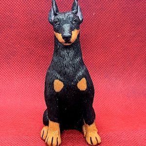 May include: A black and tan figurine of a dog sitting with its paws crossed. The dog has a short, smooth coat and a pointed muzzle.
