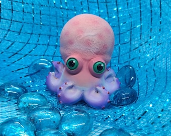 May include: A pink and purple octopus figurine with large green eyes. The octopus is sitting on a blue mesh surface with blue glass stones.