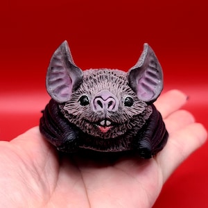 May include: A detailed figurine of a bat with a textured, gray body, large ears, and black wings. The bat has a pink nose and mouth, and is set against a red background.