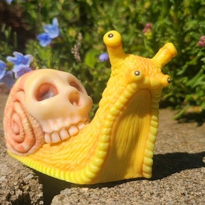 May include: A yellow and orange snail figurine with a detailed skull design. The snail has a textured shell and large, round eyes.