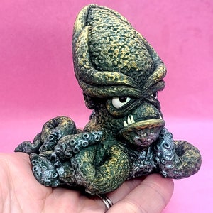 May include: A small, green and gold ceramic sculpture of an octopus with a menacing expression. The octopus has a large head with a single eye and sharp teeth. The tentacles are curled around the body.
