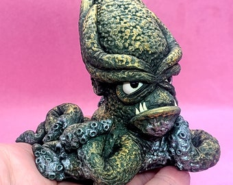 May include: A small, green and gold ceramic sculpture of an octopus with a menacing expression. The octopus has a large head with a single eye and sharp teeth. The tentacles are curled around the body.