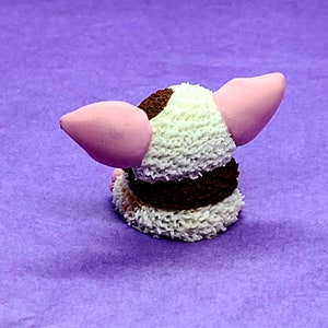 May include: A small, white and brown, fuzzy, handmade animal figurine with pink ears. The figurine is sitting on a purple surface.