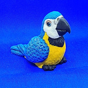 May include: A detailed figurine of a macaw parrot, featuring vibrant blue, yellow, black, and white colors. The bird is depicted in a seated position with its wings folded, showcasing intricate feather detailing and a realistic beak.