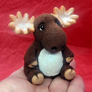 May include: A small, brown moose figurine with large, tan antlers. The moose has a white belly and is sitting on a hand.