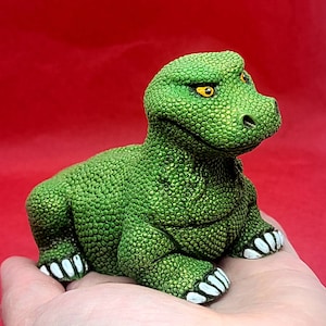 May include: A green ceramic dinosaur figurine with a textured surface and yellow eyes. The dinosaur is sitting on a hand with its mouth slightly open.