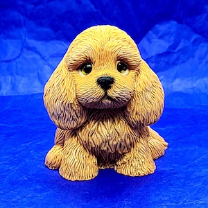 May include: A small, light brown, ceramic figurine of a dog with a fluffy coat. The dog has brown eyes and a black nose.