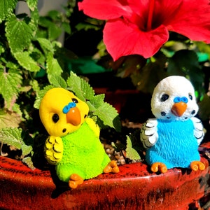 May include: Two small ceramic bird figurines, one green and yellow, the other blue and white, sitting on the edge of a red flower pot.