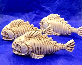 May include: Three off-white, fish-shaped sculptures with skeletal details are arranged on a blue background. The sculptures have detailed rib structures, fins, and open mouths, creating a unique decorative item.