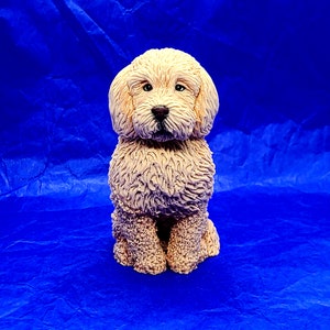 May include: A cream-colored, sculpted dog figurine with a fluffy, curly coat. The dog is sitting with its head tilted slightly to the side, looking directly at the viewer.