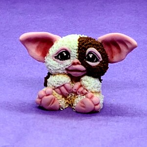 May include: A small, hand-painted figurine of a brown and white furry creature with pink ears and feet. The creature is sitting with its paws crossed and has a sweet expression on its face.