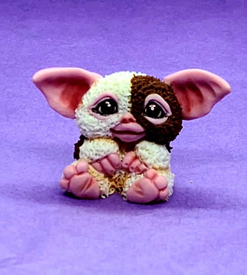 May include: A small, hand-painted figurine of a brown and white furry creature with pink ears and feet. The creature is sitting with its paws crossed and has a sweet expression on its face.