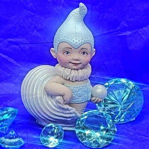 May include: A small, white porcelain figurine of a baby wearing a blue and white pointed hat and holding a pearl. The figurine is surrounded by turquoise blue, faceted gems.