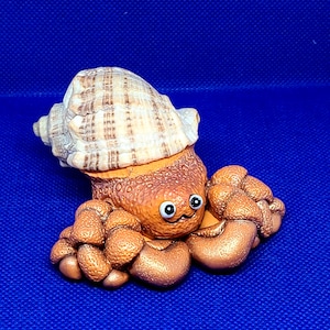 May include: A brown and gold ceramic figurine of a hermit crab with a striped shell. The crab has large, round eyes and a smiling expression.