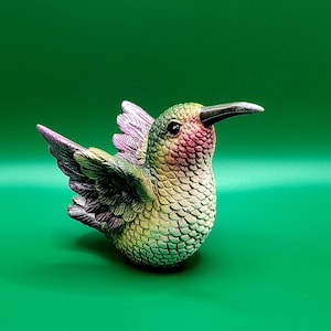 May include: A detailed figurine of a hummingbird with a green, pink, and purple color scheme. The bird has a long black beak and detailed feather textures. The figurine is set against a solid green background.