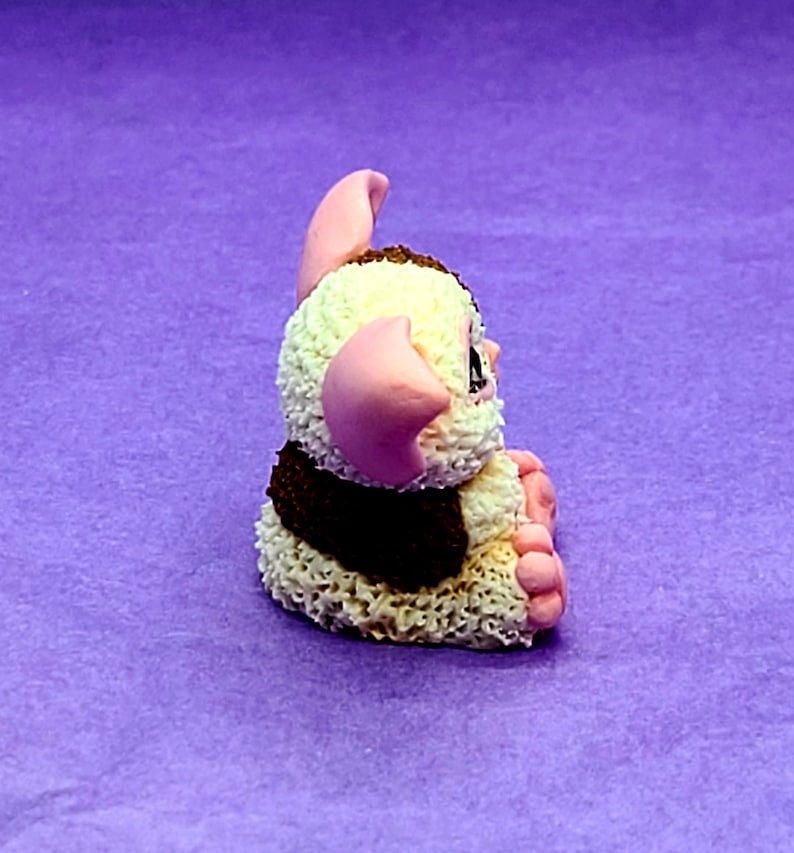 May include: A small, white and brown fuzzy toy with pink ears and feet. The toy is sitting on a purple surface.
