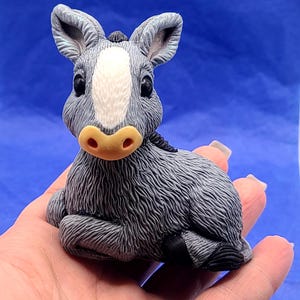 May include: A gray donkey figurine with a white stripe on its face, black eyes, and a tan nose. The donkey is in a seated position with its legs tucked in. The figurine is made of a textured material.