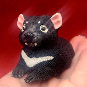 May include: A black and white figurine of a Tasmanian devil. The figurine has pink ears, white markings, and visible teeth. The devil is lying down, with its paws extended. The background is a solid red color.