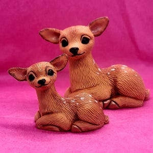 May include: Two brown ceramic deer figurines with white spots. The larger deer is sitting with its head turned to the side, while the smaller deer is sitting with its head facing forward. Both deer have black eyes and noses.