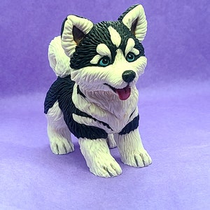 Könnte beinhalten: Eine kleine, weiße und schwarze Husky-Welpenfigur mit blauen Augen. Die Figur ist aus Polymer Clay gefertigt und steht auf einem violetten Hintergrund.