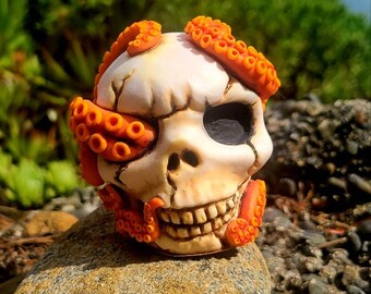 May include: A white ceramic skull with an orange octopus wrapped around it. The octopus has its tentacles covering one eye socket of the skull.
