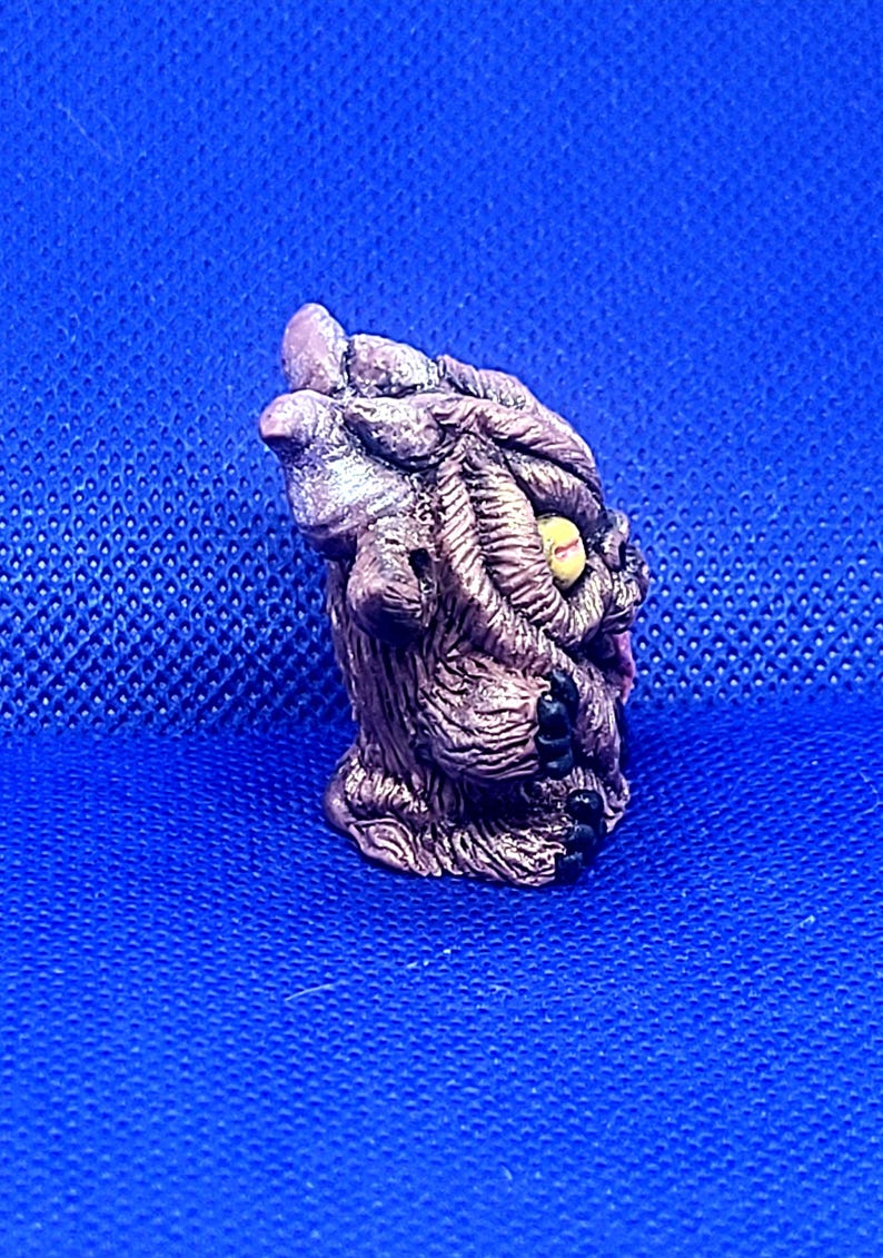 K&ouml;nnte beinhalten: Eine kleine, braune und graue, detaillierte Keramikfigur eines Wesens mit einem einzigen gelben Auge. Das Wesen hat eine strukturierte Oberfl&auml;che und scheint ein Monster oder eine mythische Kreatur zu sein.