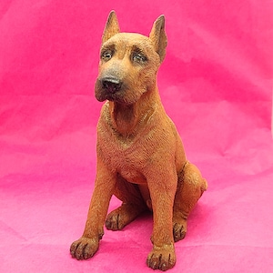 May include: A brown figurine of a dog sitting with its head tilted to the side. The dog has a short, smooth coat and a long snout.