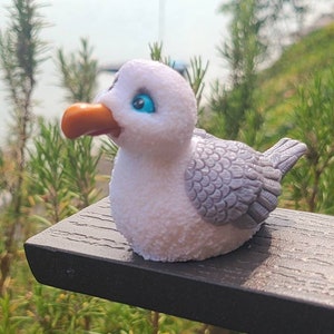 May include: A white and gray seagull figurine with blue eyes. The figurine is sitting on a dark gray wooden surface.