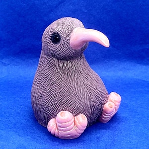 May include: A small, purple, fuzzy kiwi bird figurine with pink feet and a long, pink beak. The figurine is sitting on a blue surface.